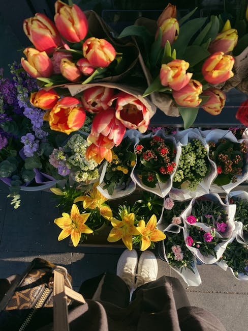 An overhead view of a display of fresh floral arrangements and bouquets at a flower shop, showing vibrant tulips in shades of orange and yellow, wrapped in brown paper, arranged on a dark surface. Sur
