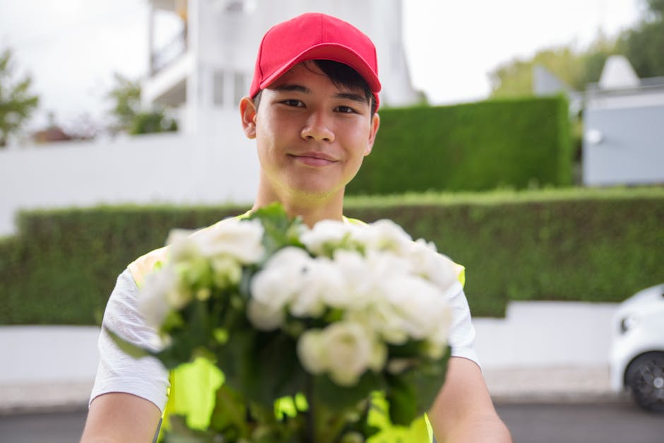 A delivery person wearing a red cap and a high-visibility vest is extending a bouquet of fresh white flowers towards the camera, with a smiling expression. The bouquet features a dense arrangement of 