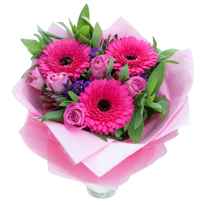 I picture this bouquet as a small, confident burst of colour that balances playfulness with quiet refinement, photographed on a clean white field so every detail reads clearly. At its heart are three vivid fuchsia gerbera daisies, each petal layered in radiating bands around a dark central disc, presenting a theatrical, joyful focal point. Around them, roses in a muted lavender-pink or dusty rose unfurl in controlled spirals, their velvety textures softening the intensity of the gerberas with romantic curves. Intermittent sprays of deep violet statice provide points of visual rest and textural contrast, like wildflower echoes tucked into a cultivated garden. The foliage is abundant and purposefully varied, with glossy, broad leaves reminiscent of salal and cooler, rounded eucalyptus tones offering depth and a faint aromatic lift. The bouquet is wrapped with tenderness: a matt pale pink tissue cushions the stems while a sheet of slightly crinkled, translucent fuchsia-pink cellophane overlays the assembly, catching ambient light and reinforcing the pink story. The top edge of a clear glass vase is visible at the base, suggesting immediacy and readiness for display on a mantle or dining table. The scent profile leans toward soft rose perfume uplifted by a green, faintly citrus leafiness. I imagine this delivered after a walk by the River Crane or left on a windowsill near Bedfont Lakes, serving as a modest but heartfelt expression of celebration, comfort, or gratitude from a florist who knows the neighbourhood well.